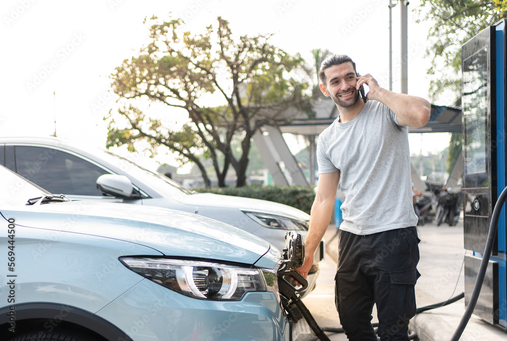 Smiling Caucasian man in casual wear Using phone while Charging on his electric car, standing on the charging station and using application. Electric car charging concept.