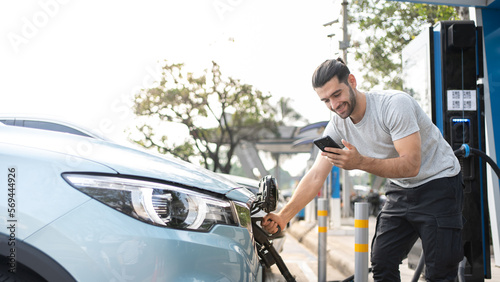 Smiling Caucasian man in casual wear Using phone while Charging on his electric car, standing on the charging station and using application. Electric car charging concept.