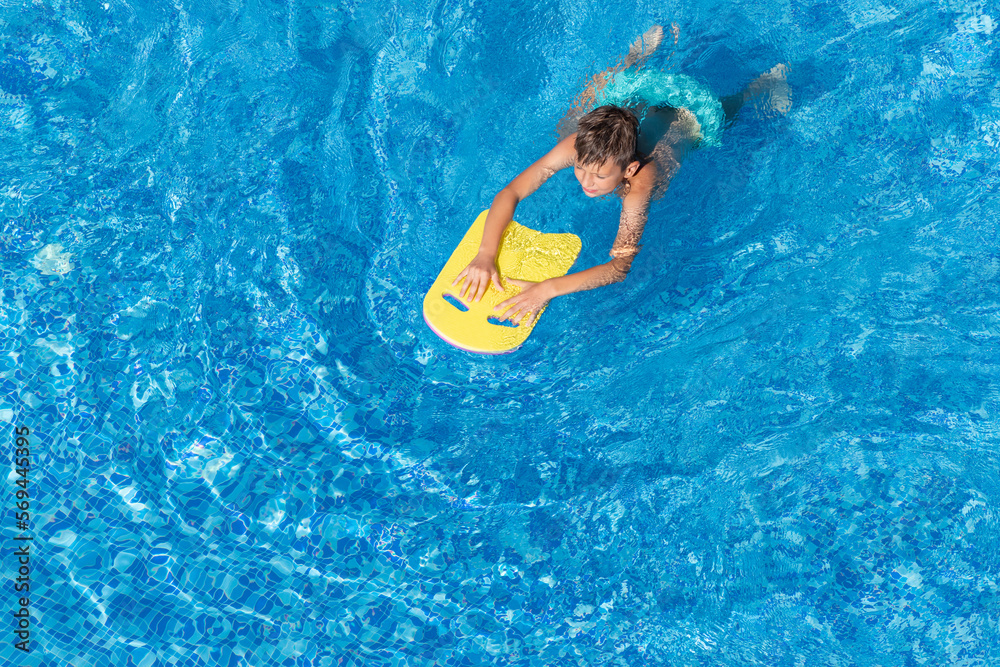 Boy child swim in swimming pool using a board. Active wellness summer vacation in resort hotel. Water sports and game for children on summer holiday. View from above