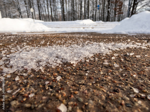 Salt grains on icy sidewalk surface in the winter. Applying salt to keep roads clear and people safe in winter weather from ice or snow. Macro view of salt grains with winter scenery in bacground