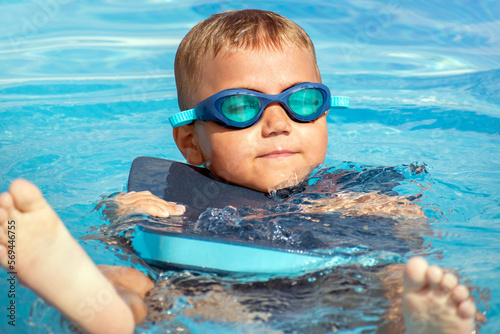 Boy child in swimming goggles with float foam board tool swim in water pool. Safe pool training, summer holiday, family vacation, travel, water sport concept.