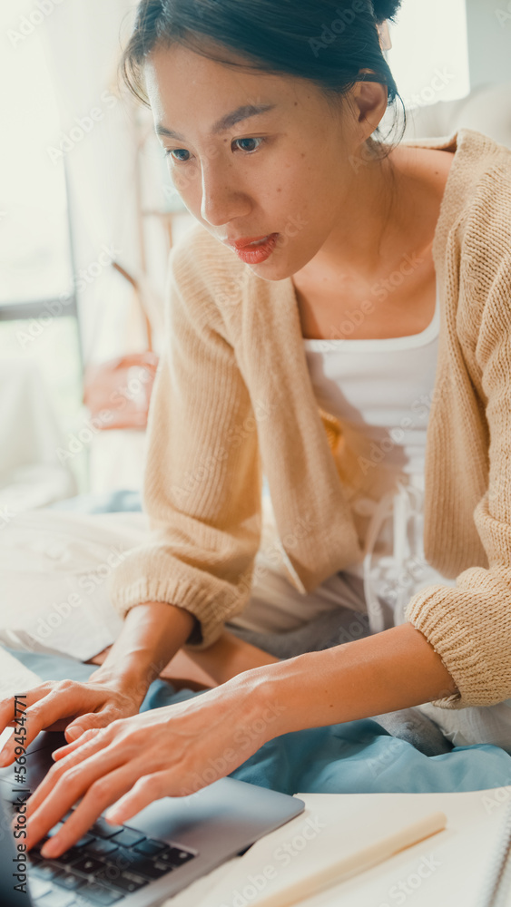 Happy young Asian girl with white cream cardigan on bed focus computer laptop full of paperwork messy document work idea in cozy bedroom at home in morning. Work from home concept. Vertical Screen. Happy young Asian girl with white cream cardigan on bed focus computer laptop full of paperwork messy document work idea in cozy bedroom at home in morning. Work from home concept. Vertical Screen.