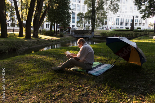 An elderly man in a white shirt is sitting on a blanket, on the ground in a park and reading an interesting book. A pensioner alone is resting in nature, passionate about his hobby