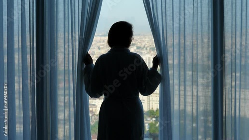 Woman comes to the window, carefully unveils transparent curtains and looks incredulously at city. Long quarantine or self-isolation is bad for the mental and physical health of people photo