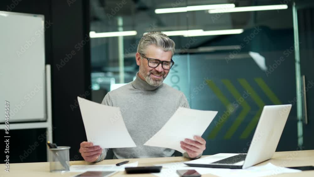 Smiling mature gray haired bearded businessman looking through business papers at desk at workplace in modern office. The happy entrepreneur is satisfied with what he has read and signs the documents Smiling mature gray haired bearded businessman looking through business papers at desk at workplace in modern office. The happy entrepreneur is satisfied with what he has read and signs the documents