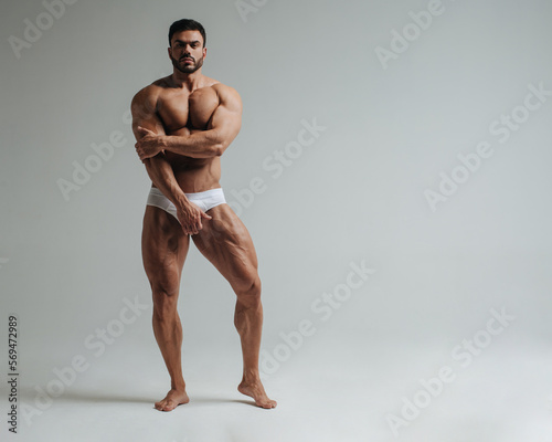 Sexy muscular man in white underwear posing in studio. Handsome bodybuilder standing on grey background. Big male model showing his muscles.