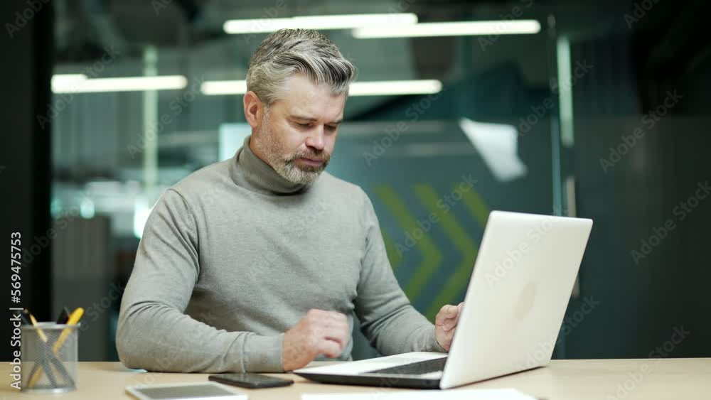Confused mature gray haired bearded businessman is solving a complex problem while working on a laptop while sitting at a desk in modern office. Disappointed entrepreneur can't find the right solution Confused mature gray haired bearded businessman is solving a complex problem while working on a laptop while sitting at a desk in modern office. Disappointed entrepreneur can't find the right solution