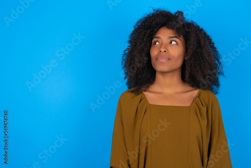 young woman with afro hairstyle wearing brown dress over blue wall looking aside into empty space thoughtful © Jihan