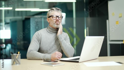 Wallpaper Mural Mature gray haired bearded businessman wearing glasses has a toothache while sitting at a desk at a workplace in modern office. Tired middle aged entrepreneur holding hand on cheek massaging sore spot Torontodigital.ca