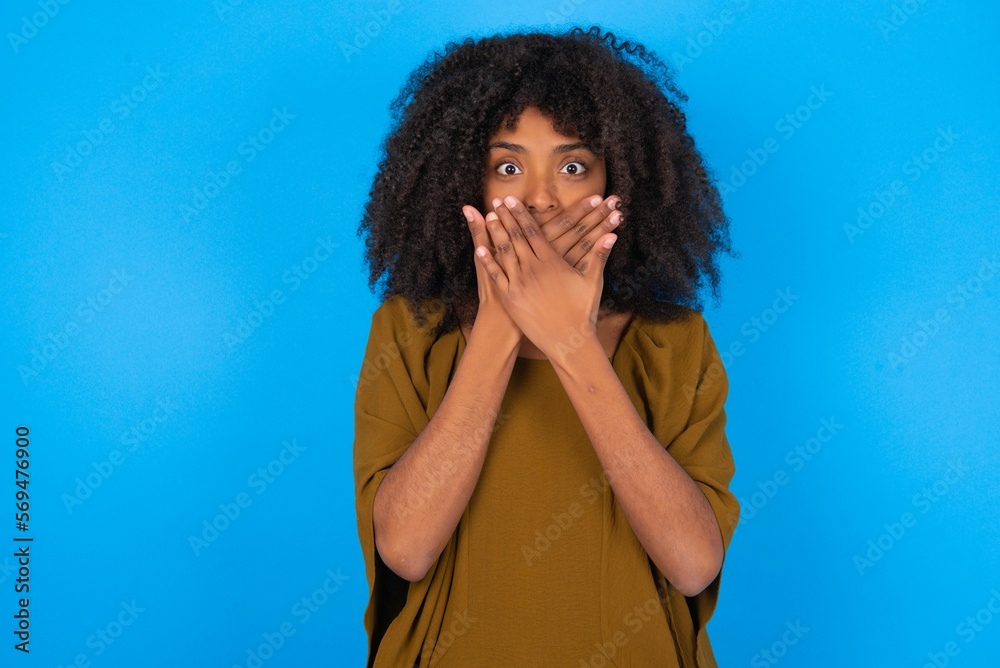 Φωτογραφία young woman with afro hairstyle wearing brown dress over blue wall shocked covering mouth with hands for mistake