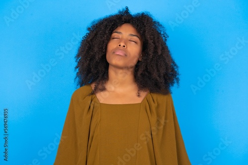 young woman with afro hairstyle wearing brown dress over blue wall looking sleepy and tired, exhausted for fatigue and hangover, lazy eyes in the morning. © Jihan