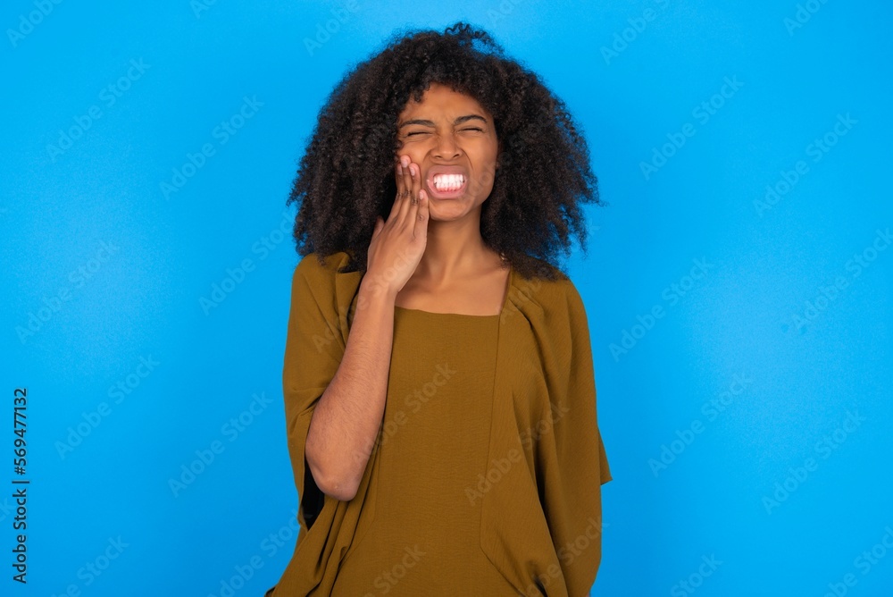 young woman with afro hairstyle wearing brown dress over blue wall touching mouth with hand with painful expression because of toothache or dental illness on teeth. young woman with afro hairstyle wearing brown dress over blue wall touching mouth with hand with painful expression because of toothache or dental illness on teeth.