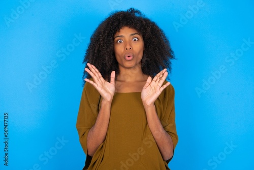young woman with afro hairstyle wearing brown dress over blue wall Moving away hands palms showing refusal and denial with afraid and disgusting expression. Stop and forbidden. © Jihan
