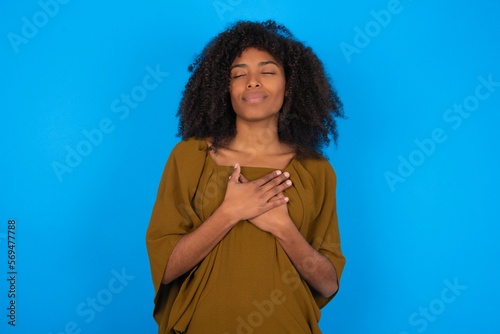 young woman with afro hairstyle wearing brown dress over blue wall smiling with hands on chest with closed eyes and grateful gesture on face. Health concept.