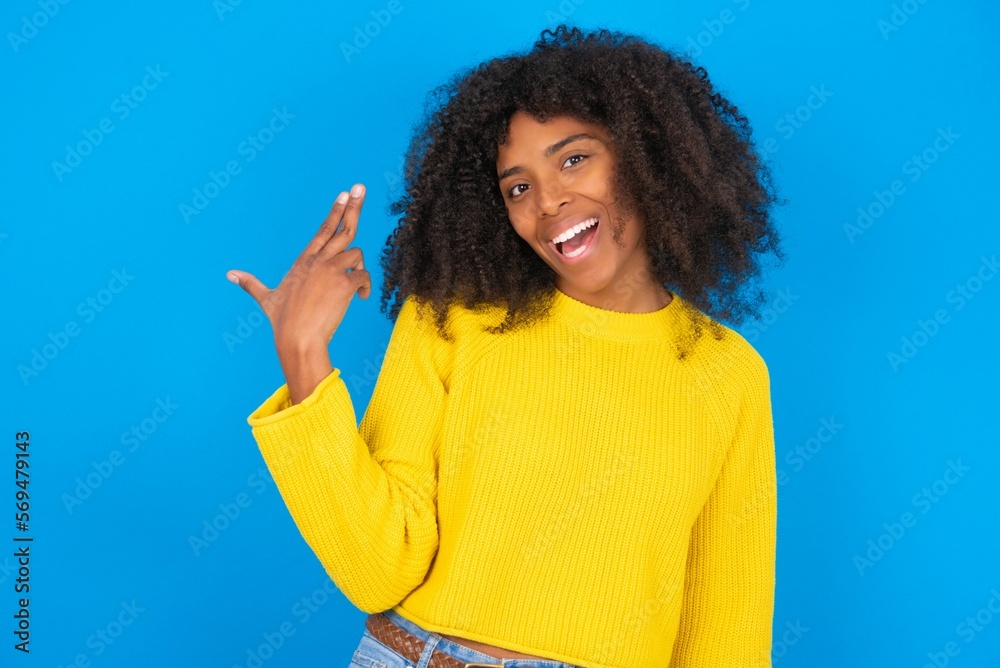 young woman with afro hairstyle wearing yellow sweater over blue wall foolishness around shoots in temple with fingers makes suicide gesture. Funny model makes finger gun pistol young woman with afro hairstyle wearing yellow sweater over blue wall foolishness around shoots in temple with fingers makes suicide gesture. Funny model makes finger gun pistol