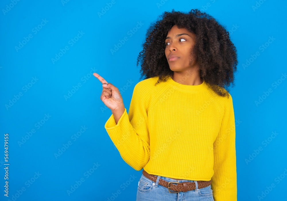 young woman with afro hairstyle wearing yellow sweater over blue wall points at copy space and advertises something, advices best price. young woman with afro hairstyle wearing yellow sweater over blue wall points at copy space and advertises something, advices best price.
