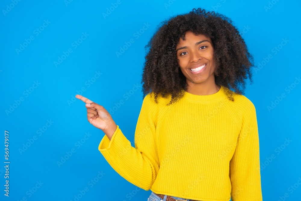 young woman with afro hairstyle wearing yellow sweater over blue wall points aside on copy blank space. People promotion and advertising concept young woman with afro hairstyle wearing yellow sweater over blue wall points aside on copy blank space. People promotion and advertising concept