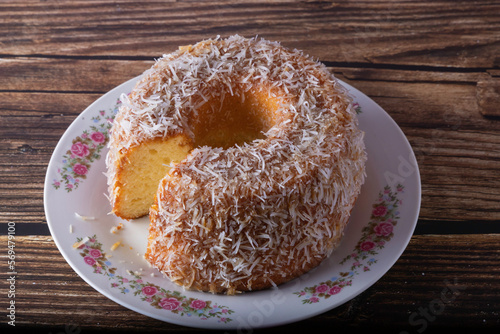 Coconut-flavored sweet cake topped with coconut flakes, with a cut slice showing the delicious dough on a decorated plate. Using wooden board and wooden background. Selective focus.