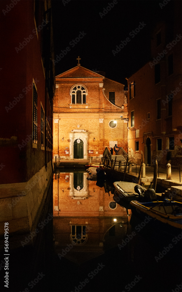 Venice landscape reflections. San Martino church reflected in canal water at night in winter. Venice, Italy. Silhouettes of couple in masks on the bridge returning from Carnival events. 
