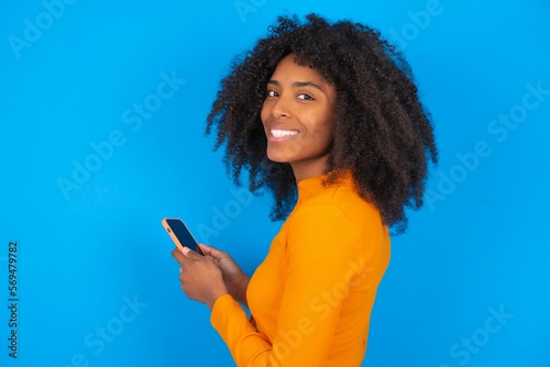 Rear view photo portrait of young beautiful brunette woman wearing colourful dress over white wall using smartphone smiling