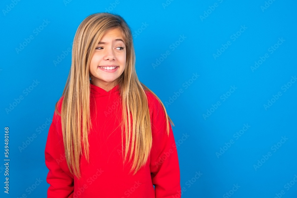 Oops! Portrait of caucasian teen girl wearing red sweater over blue studio background clenches teeth and looks confusedly aside, realizes her bad mistake, Oops! Portrait of caucasian teen girl wearing red sweater over blue studio background clenches teeth and looks confusedly aside, realizes her bad mistake,
