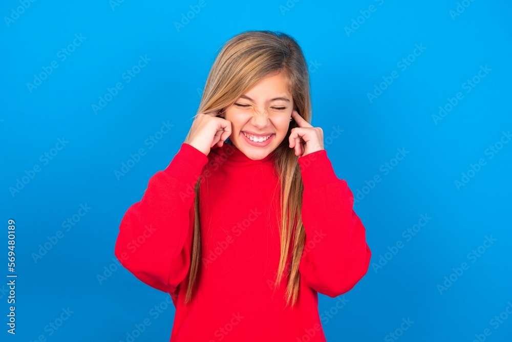 Stop making this annoying sound! Unhappy stressed out caucasian teen girl wearing red sweater over blue studio background making worry face, plugging ears with fingers, irritated with loud noise. Stop making this annoying sound! Unhappy stressed out caucasian teen girl wearing red sweater over blue studio background making worry face, plugging ears with fingers, irritated with loud noise.