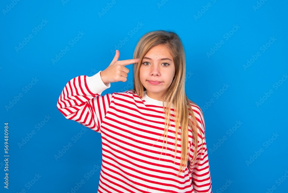 Unhappy caucasian teen girl wearing striped shirt over blue studio background curves lips and makes suicide gesture, shoots in temple with hand, tries kill himself. Unhappy caucasian teen girl wearing striped shirt over blue studio background curves lips and makes suicide gesture, shoots in temple with hand, tries kill himself.