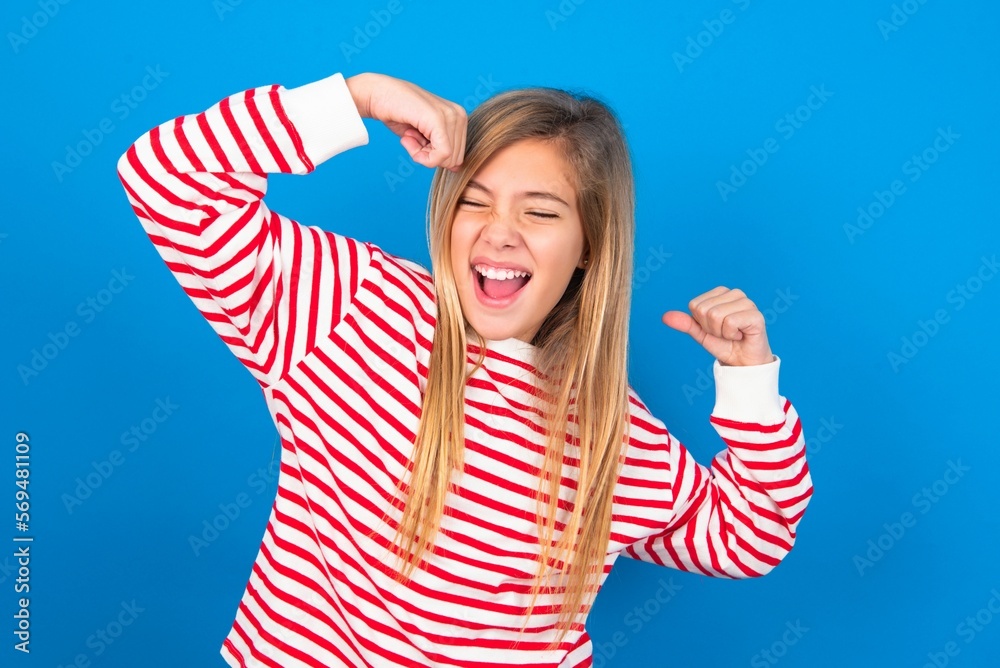 Yes I am winner. Portrait of charming delighted and excited caucasian teen girl wearing striped shirt over blue raising up fist in triumph and victory smiling achieving success grinning from delight. Yes I am winner. Portrait of charming delighted and excited caucasian teen girl wearing striped shirt over blue raising up fist in triumph and victory smiling achieving success grinning from delight.