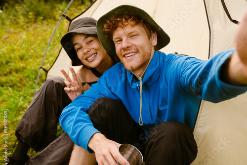 Joyful tourist couple taking selfie while sitting at camping tent in forest