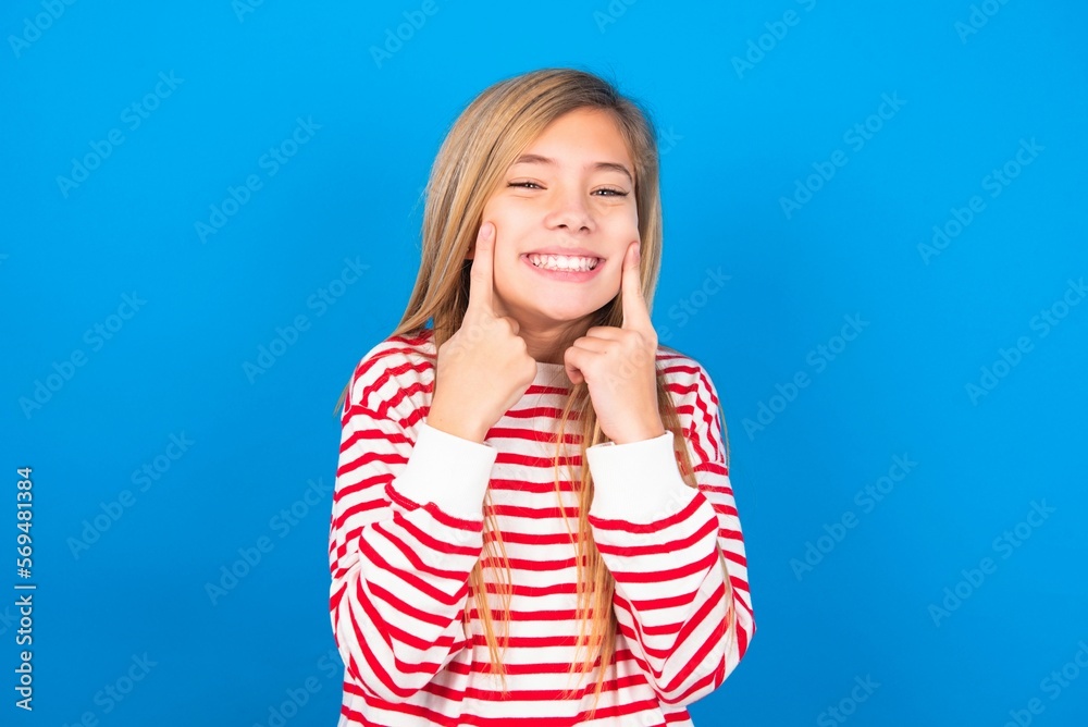 Strong healthy straight white teeth. Close up portrait of happy caucasian teen girl wearing striped shirt over blue studio background with beaming smile pointing on perfect clear white teeth. Strong healthy straight white teeth. Close up portrait of happy caucasian teen girl wearing striped shirt over blue studio background with beaming smile pointing on perfect clear white teeth.