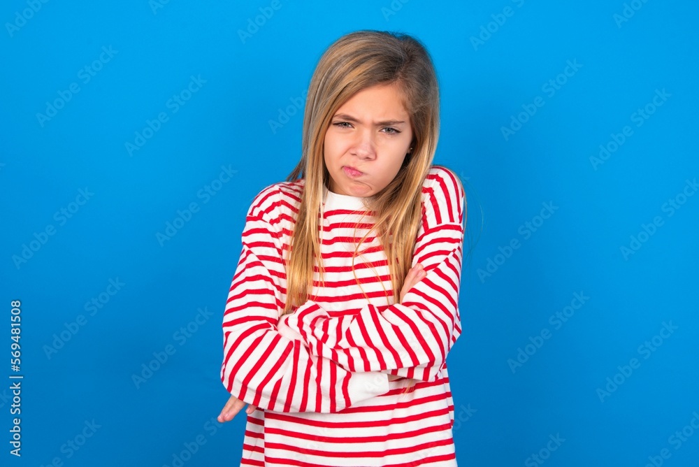 Picture of angry caucasian teen girl wearing striped shirt over blue studio background crossing arms. Looking at camera with disappointed expression. Picture of angry caucasian teen girl wearing striped shirt over blue studio background crossing arms. Looking at camera with disappointed expression.