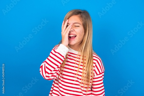 Charismatic carefree joyful caucasian teen girl wearing striped shirt over blue studio background likes laugh out loud not hiding emotions giggling hear funny hilarious joke chuckling facepalm.
