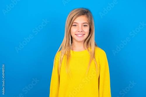 caucasian teen girl wearing yellow sweater over blue studio background  with nice beaming smile pleased expression. Positive emotions concept photo