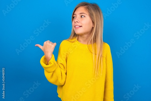 caucasian teen girl wearing yellow sweater over blue studio background points away and gives advice demonstrates advertisement