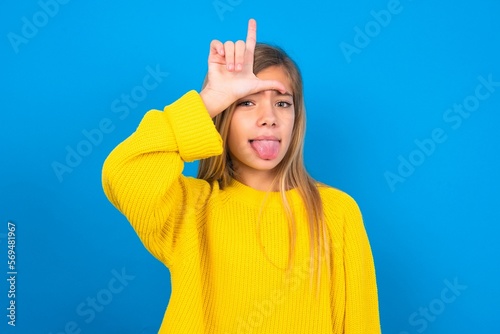 Funny caucasian teen girl wearing yellow sweater over blue studio background makes loser gesture mocking at someone sticks out tongue making grimace face.