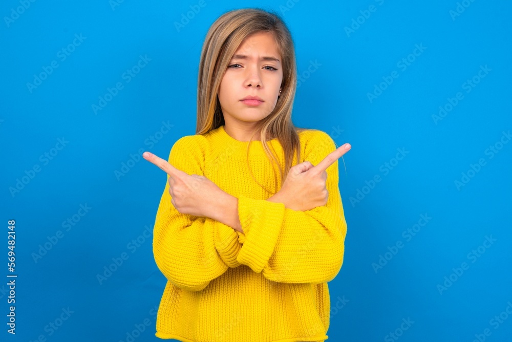 Serious caucasian teen girl wearing yellow sweater over blue studio background crosses hands and points at different sides hesitates between two items. Hard decision concept