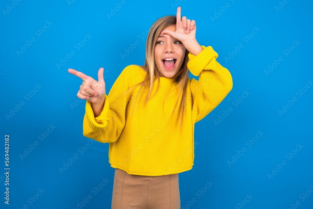 caucasian teen girl wearing yellow sweater over blue studio background showing loser sign and pointing at empty space caucasian teen girl wearing yellow sweater over blue studio background showing loser sign and pointing at empty space
