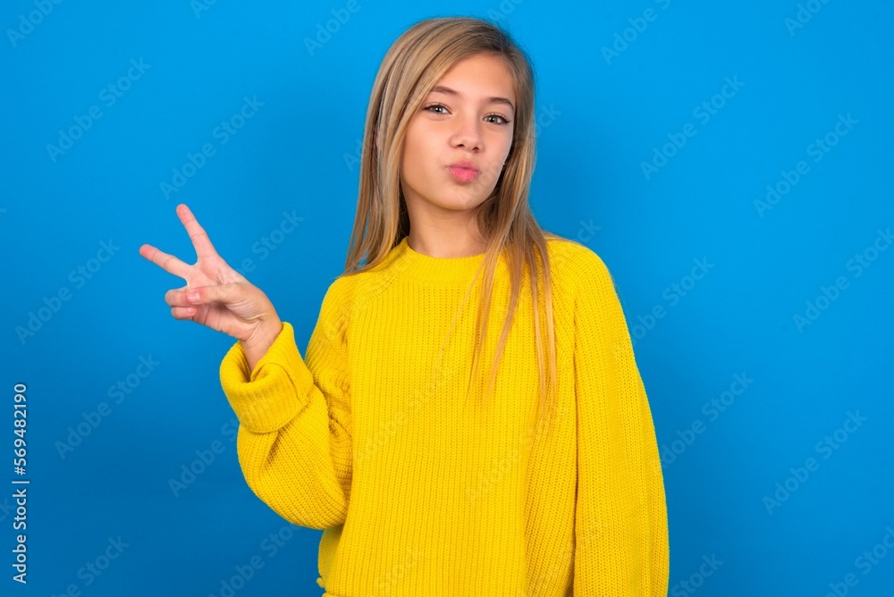 caucasian teen girl wearing yellow sweater over blue studio background makes peace gesture keeps lips folded shows v sign. Body language concept