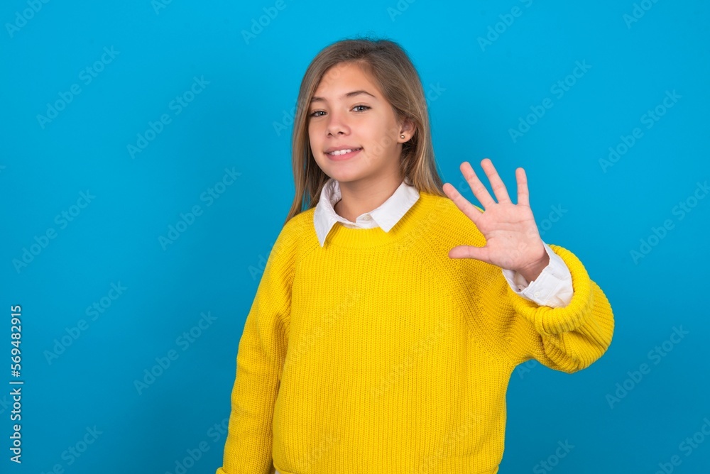 caucasian teen girl wearing yellow sweater over blue studio background Waiving saying hello happy and smiling, friendly welcome gesture.