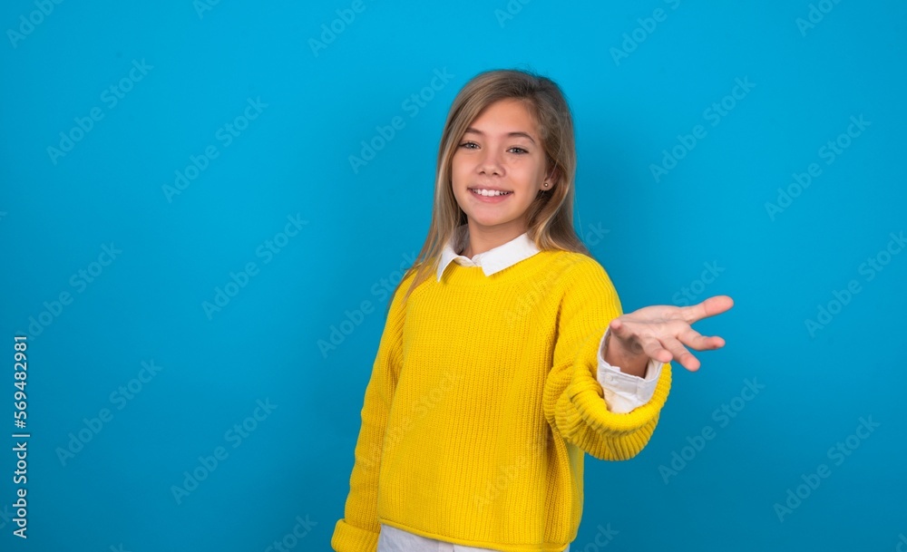 caucasian teen girl wearing yellow sweater over blue studio background smiling cheerful offering palm hand giving assistance and acceptance. caucasian teen girl wearing yellow sweater over blue studio background smiling cheerful offering palm hand giving assistance and acceptance.