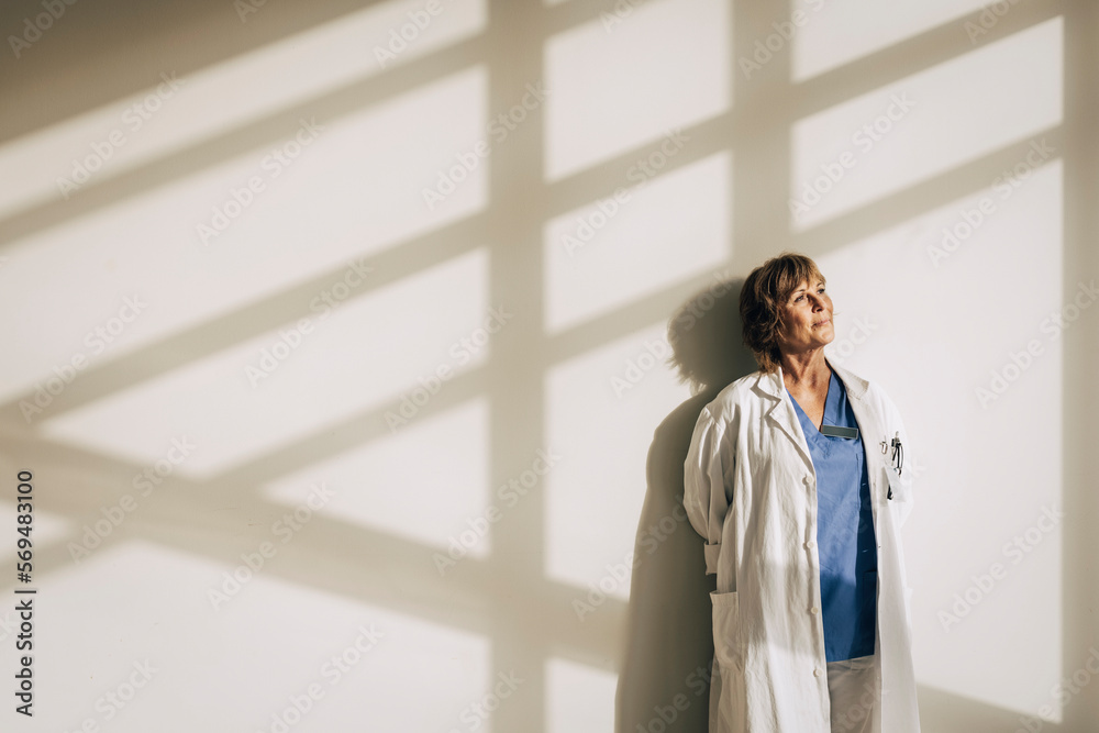 Female doctor wearing lab coat day dreaming while standing against wall with sunlight Female doctor wearing lab coat day dreaming while standing against wall with sunlight