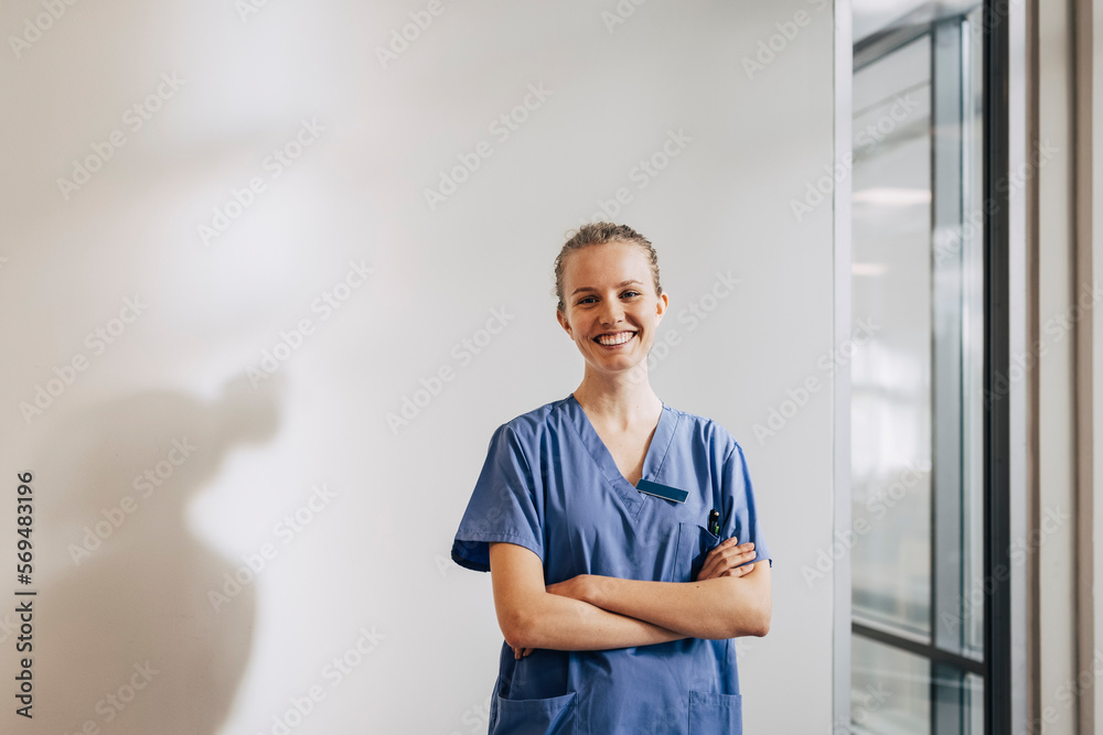 Portrait of happy young female nurse standing with arms crossed against wall at hospital