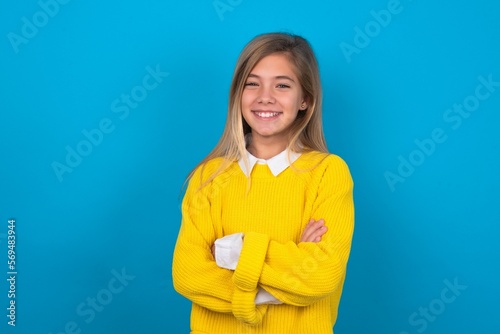 caucasian teen girl wearing yellow sweater over blue studio background happy face smiling with crossed arms looking at the camera. Positive person.