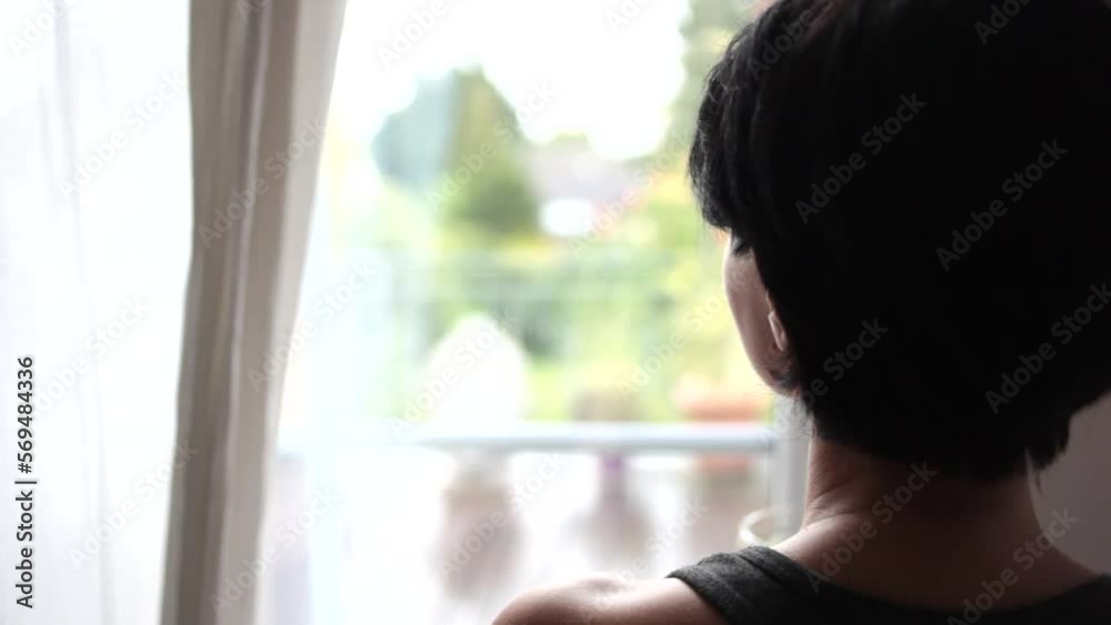 Handheld shot of mature woman holding green tea in cup while looking through window at home