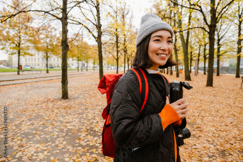 Smiling young asian woman drinking hot tea from thermos during walk in park