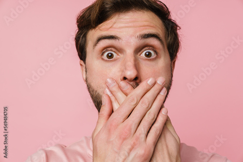 Shocked man with wide opened eyes covering his mouth with hands while standing isolated