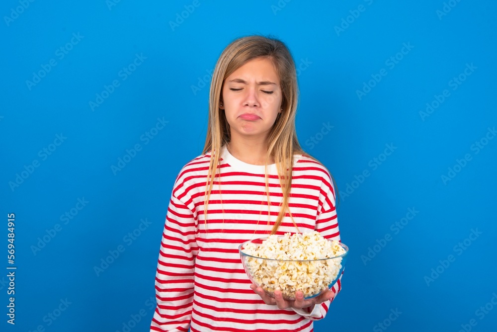 Dismal gloomy rejected beautiful caucasian teen girl wearing striped T-shirt over blue wall eating popcorn has problems and difficulties, curves lower lip in despair, being in depression