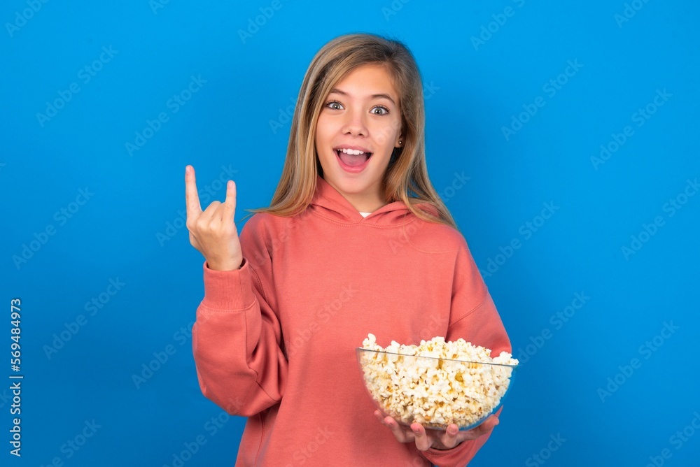 beautiful caucasian teen girl wearing pink sweater over blue wall eating popcorn makes rock n roll sign looks self confident and cheerful enjoys cool music at party. Body language concept. beautiful caucasian teen girl wearing pink sweater over blue wall eating popcorn makes rock n roll sign looks self confident and cheerful enjoys cool music at party. Body language concept.