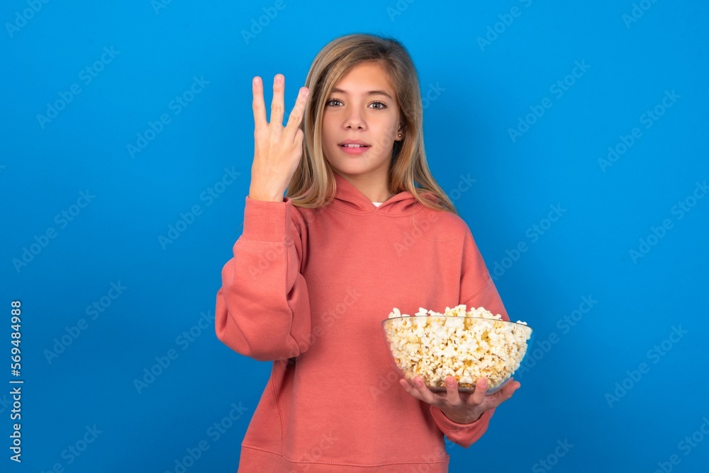 beautiful caucasian teen girl wearing pink sweater over blue wall eating popcorn smiling and looking friendly, showing number three or third with hand forward, counting down beautiful caucasian teen girl wearing pink sweater over blue wall eating popcorn smiling and looking friendly, showing number three or third with hand forward, counting down