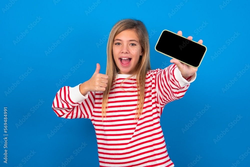 Portrait beautiful caucasian teen girl wearing striped T-shirt over blue wall holding in hands cell showing giving black screen thumb up Portrait beautiful caucasian teen girl wearing striped T-shirt over blue wall holding in hands cell showing giving black screen thumb up